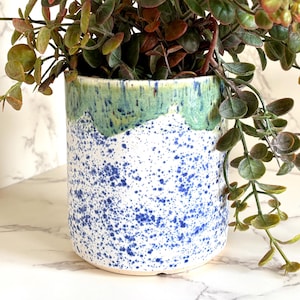 May include: A cylindrical ceramic planter with a white base and blue splatter design. The top rim is painted with a green and blue glaze. The planter holds a trailing green and brown plant, creating a decorative display.