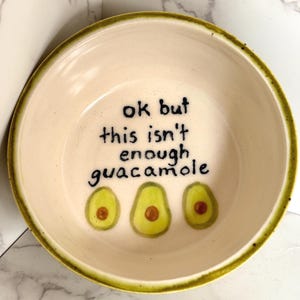 May include: A cream-colored ceramic bowl with a green rim and the text "ok but this isn't enough guacamole" in black. Three avocado halves are painted below the text.