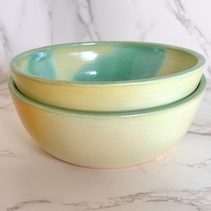 May include: Two ceramic bowls stacked on top of each other. The bowls are a pale yellow color with a teal and yellow interior. The bowls have a smooth, glossy finish and are likely handmade.