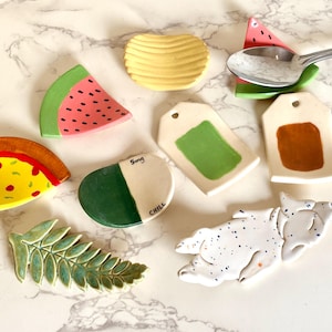 May include: A collection of whimsical ceramic spoon rests and trinket dishes in various shapes, including watermelon slices, pizza, a potato chip, and a dog. The pieces are hand-painted with bright colors and playful designs.
