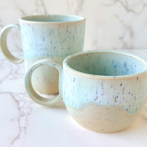 May include: Two handmade ceramic mugs. The larger mug has a handle and a cylindrical shape. The smaller mug is a rounded cup. Both mugs are light blue with speckled detailing and a cream-colored base.