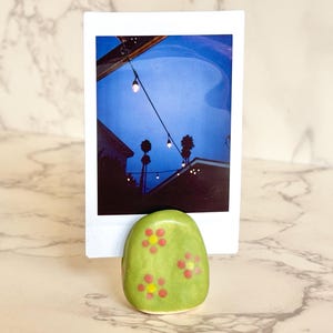 May include: A green ceramic photo holder with red and yellow flower designs. The holder is holding a polaroid photo of a night scene with string lights and a blue sky. The holder is on a marble surface.