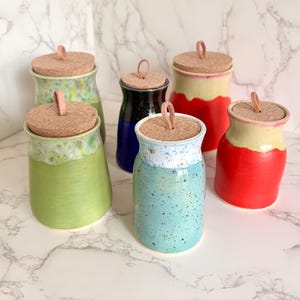 May include: A collection of six ceramic jars with cork lids. The jars are in various colors, including green, blue, red, and black. Each jar has a leather loop attached to the cork for easy removal. The jars are displayed on a white marble surface.