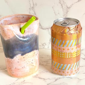 Puede incluir: Un vaso de cer&aacute;mica con una pajita verde y una lata decorada. El vaso tiene una combinaci&oacute;n de colores rosa, azul y blanco. La lata est&aacute; envuelta en cinta decorativa dorada y rosa estampada.