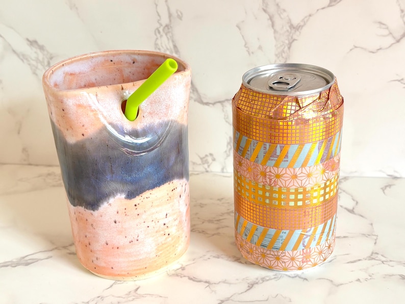 Puede incluir: Un vaso de cer&aacute;mica con una pajita verde y una lata decorada. El vaso tiene una combinaci&oacute;n de colores rosa, azul y blanco. La lata est&aacute; envuelta en cinta decorativa dorada y rosa estampada.