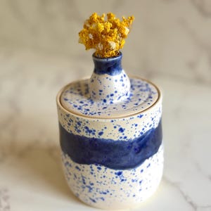May include: A cylindrical ceramic jar with a lid featuring a small central opening. The surface is off-white with blue paint splatters and a wide, horizontal dark blue band. A small bouquet of dried yellow flowers is placed in the lid opening.