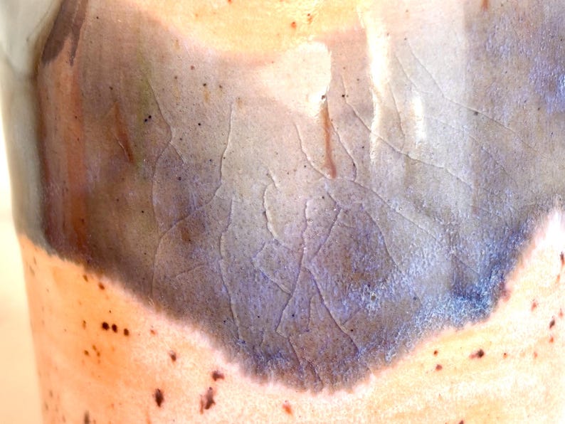 Puede incluir: Primer plano de un recipiente de cer&aacute;mica con una superficie texturizada. El esmalte transita de un melocot&oacute;n claro a un azul-gris oscuro, con patrones de craquelado visibles. Peque&ntilde;os puntos oscuros est&aacute;n dispersos por la superficie.