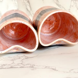 Puede incluir: Dos macetas de cer&aacute;mica hechas a mano. Las macetas son de color rosa claro con una franja gris oscuro y un interior naranja moteado. Cada maceta tiene una abertura curva &uacute;nica y un peque&ntilde;o agujero en la base.