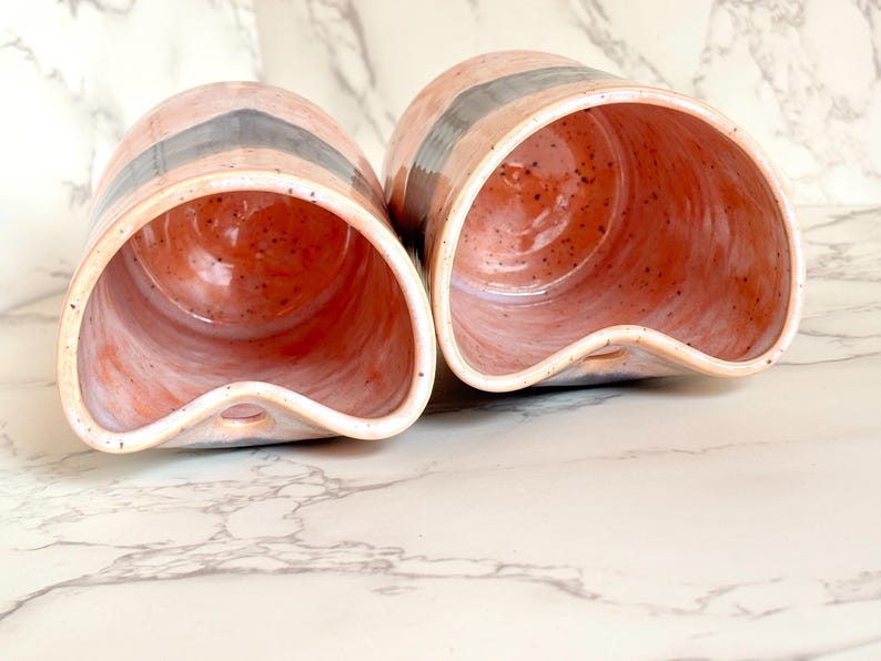 Puede incluir: Dos macetas de cer&aacute;mica hechas a mano. Las macetas son de color rosa claro con una franja gris oscuro y un interior naranja moteado. Cada maceta tiene una abertura curva &uacute;nica y un peque&ntilde;o agujero en la base.