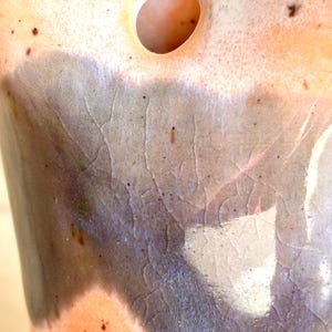 Puede incluir: Primer plano de un jarr&oacute;n de cer&aacute;mica con una superficie texturizada &uacute;nica. El jarr&oacute;n presenta una mezcla de colores, incluyendo marr&oacute;n, gris y naranja, con un patr&oacute;n craquelado. Se ve una abertura circular en la parte superior.