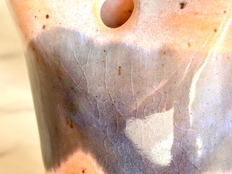 Puede incluir: Primer plano de un jarr&oacute;n de cer&aacute;mica con una superficie texturizada &uacute;nica. El jarr&oacute;n presenta una mezcla de colores, incluyendo marr&oacute;n, gris y naranja, con un patr&oacute;n craquelado. Se ve una abertura circular en la parte superior.