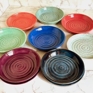May include: A collection of eight ceramic bowls in various colors, including red, green, blue, and purple. Each bowl has a spiral design in the center. The bowls are arranged on a white marble surface.