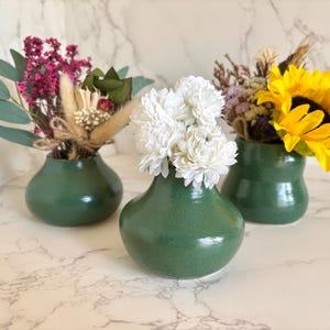 May include: Three small, green ceramic vases each hold a different arrangement of flowers. One vase has white flowers, another has a yellow sunflower, and the third has a mix of dried flowers and greenery.
