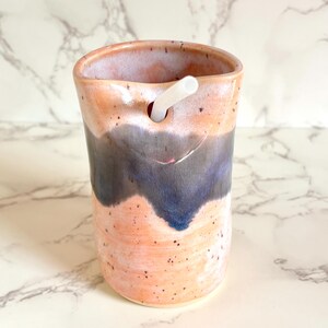 Puede incluir: Vaso de cer&aacute;mica hecho a mano con un dise&ntilde;o &uacute;nico. El vaso presenta una combinaci&oacute;n de colores melocot&oacute;n y azul con una pajita blanca insertada. La taza de cer&aacute;mica tiene una textura moteada y un dise&ntilde;o ondulado.
