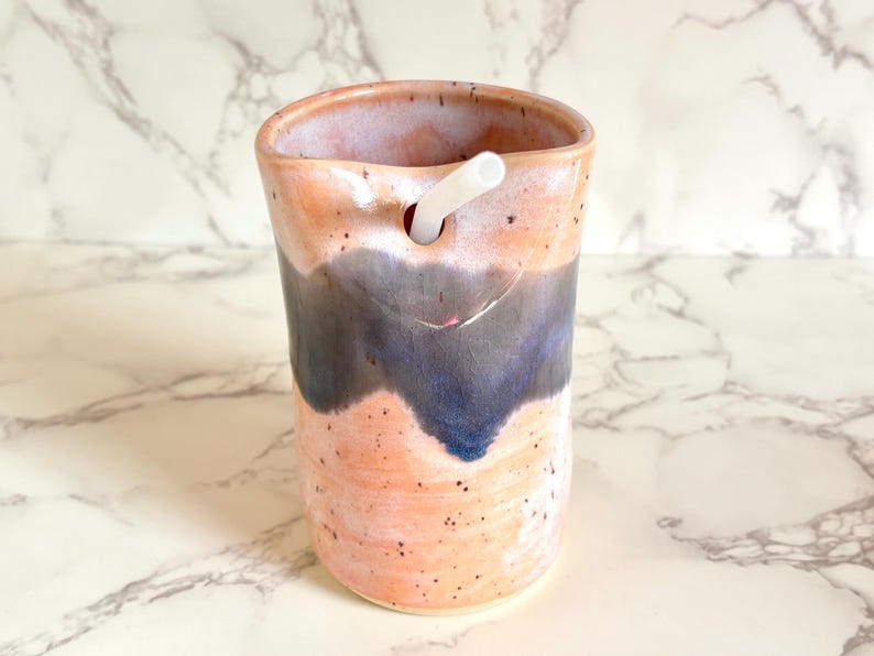 Puede incluir: Vaso de cer&aacute;mica hecho a mano con un dise&ntilde;o &uacute;nico. El vaso presenta una combinaci&oacute;n de colores melocot&oacute;n y azul con una pajita blanca insertada. La taza de cer&aacute;mica tiene una textura moteada y un dise&ntilde;o ondulado.