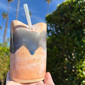 Puede incluir: Un vaso de cer&aacute;mica hecho a mano con un dise&ntilde;o &uacute;nico. La taza presenta una base de color naranja claro con un patr&oacute;n de ondas azul oscuro y gris cerca de la parte superior. Se inserta una pajita transparente. El fondo muestra palmeras y vegetaci&oacute;n.