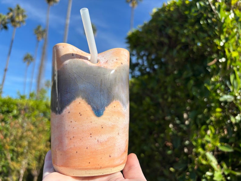Puede incluir: Un vaso de cer&aacute;mica hecho a mano con un dise&ntilde;o &uacute;nico. La taza presenta una base de color naranja claro con un patr&oacute;n de ondas azul oscuro y gris cerca de la parte superior. Se inserta una pajita transparente. El fondo muestra palmeras y vegetaci&oacute;n.