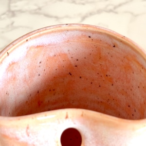 Puede incluir: Un bolsillo de pared de cer&aacute;mica hecho a mano en color melocot&oacute;n con una textura moteada. El interior es de un tono melocot&oacute;n m&aacute;s claro con un borde blanco. Un peque&ntilde;o agujero redondo es visible en la parte inferior. Ideal para plantas o objetos peque&ntilde;os.