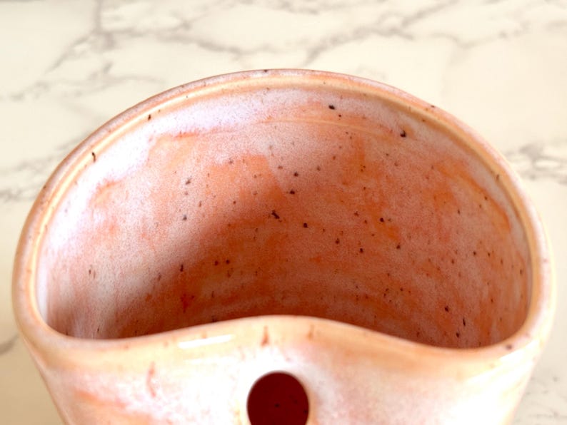 Puede incluir: Un bolsillo de pared de cer&aacute;mica hecho a mano en color melocot&oacute;n con una textura moteada. El interior es de un tono melocot&oacute;n m&aacute;s claro con un borde blanco. Un peque&ntilde;o agujero redondo es visible en la parte inferior. Ideal para plantas o objetos peque&ntilde;os.