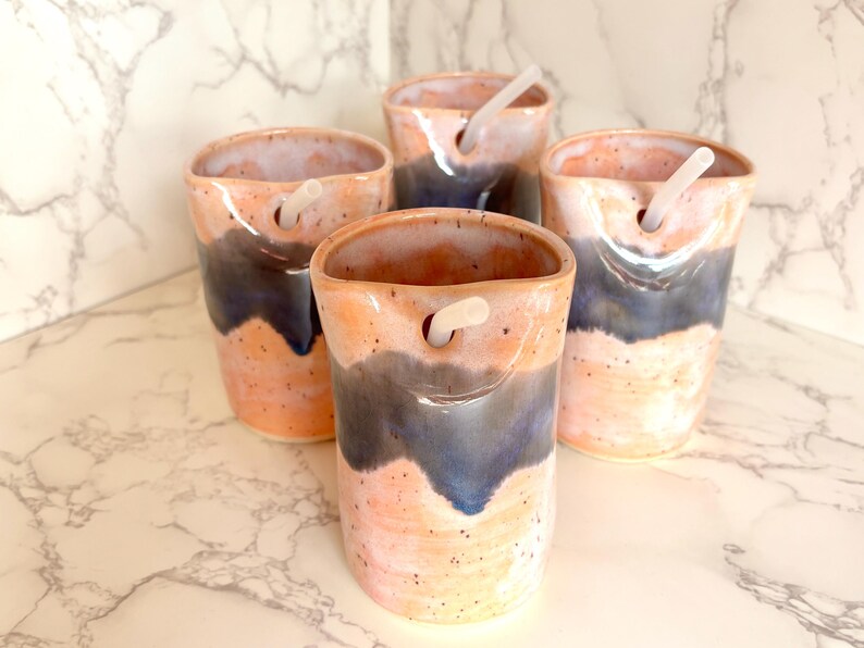 Puede incluir: Cuatro vasos de cer&aacute;mica hechos a mano con un dise&ntilde;o &uacute;nico. Las tazas presentan una combinaci&oacute;n de colores melocot&oacute;n y azul con una textura moteada. Cada vaso tiene un agujero cerca de la parte superior e incluye una pajita blanca.