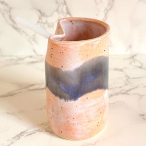 Puede incluir: Un vaso de cer&aacute;mica hecho a mano con un dise&ntilde;o &uacute;nico. El vaso presenta un exterior color melocot&oacute;n con un patr&oacute;n de ondas azul oscuro y gris. Una pajita blanca descansa en la abertura. Ideal para bebidas.