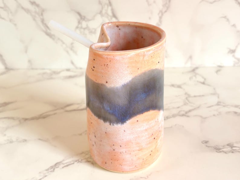Puede incluir: Un vaso de cer&aacute;mica hecho a mano con un dise&ntilde;o &uacute;nico. El vaso presenta un exterior color melocot&oacute;n con un patr&oacute;n de ondas azul oscuro y gris. Una pajita blanca descansa en la abertura. Ideal para bebidas.