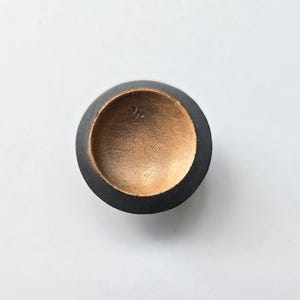 May include: A circular knob featuring a matte black outer rim and a recessed, metallic gold-toned centre. The interior of the gold-coloured basin displays a smooth, slightly textured surface with a subtle, circular indentation at the very bottom.