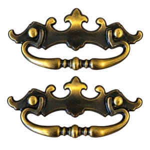 May include: Two matching drawer pulls featuring an ornate, symmetrical backplate with a dark, antiqued brass finish. Each pull includes a curved, swinging handle attached to the decorative mounting plate, showcasing a polished gold-tone metal.
