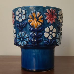 May include: A cylindrical ceramic planter with a deep blue glazed finish and a textured wood-grain surface. The exterior features raised, embossed floral motifs in white, orange, and pink, arranged in a repeating pattern around the upper section.
