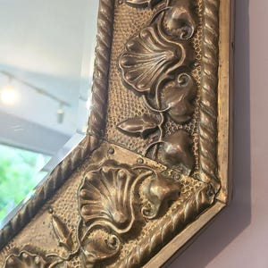 May include: Ornate, antique mirror with a decorative, gold-colored frame. The frame features a textured surface with floral and leaf motifs, and a rope-like border. The mirror's shape is a modified octagon.