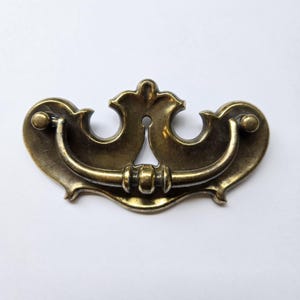 Peut inclure: Une poignée de tiroir en métal couleur laiton dotée d'une plaque arrière décorative aux bords incurvés et symétriques avec une découpe centrale. Une poignée incurvée et articulée est fixée à la plaque, avec un détail central perlé.