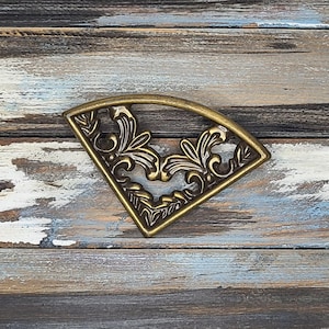 May include: A decorative, fan-shaped metal piece with an ornate, floral design. The piece is made of a gold-colored metal and features intricate details. The design includes leaves and swirling patterns, creating a vintage aesthetic.
