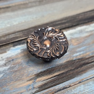 May include: A round, ornate, copper-toned cabinet knob with a floral design.