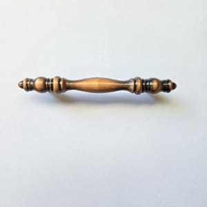 May include: A horizontal cabinet pull handle featuring a metallic copper finish with dark, antiqued accents. The handle has a smooth, rounded centre section that tapers towards both ends, which are decorated with a series of raised, ringed details.