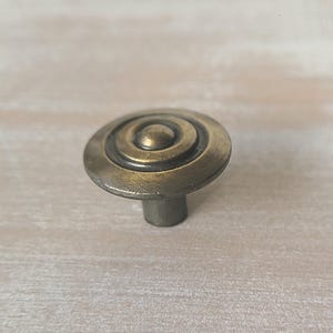 Peut inclure: Un bouton de meuble de couleur bronze avec un motif circulaire. Le bouton présente des anneaux concentriques autour d'un cercle central surélevé et une courte tige cylindrique. La quincaillerie a une esthétique vintage ou antique.