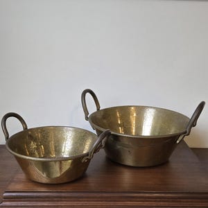 May include: Two round brass bowls with a polished finish and a slightly weathered patina. Each bowl features two curved metal handles attached to the sides. The bowls are of different sizes, with the larger one positioned behind the smaller one.