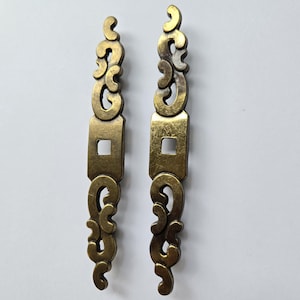 May include: Two elongated, vertical metal drawer pulls with an antique brass finish. Each piece features a central rectangular section with a square hole and symmetrical, openwork scroll patterns extending from the top and bottom ends.