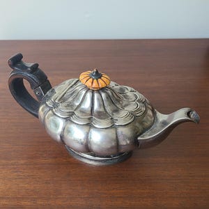 May include: A silver-colored teapot with a black handle and a decorative lid. The lid features a small, orange and yellow finial. The teapot has a bulbous body and a spout for pouring. The teapot sits on a round base.