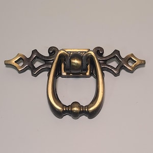 May include: A vintage-style brass drawer pull with a decorative design. The pull features a curved handle and ornate detailing on the backplate, with diamond-shaped cutouts on each end. The metal has a warm, aged finish.