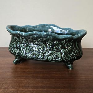 May include: A deep teal ceramic planter shaped like a miniature clawfoot bathtub. The exterior features a raised, intricate floral and scroll relief pattern with a glossy finish. The rim is wavy, and the vessel stands on four small feet.