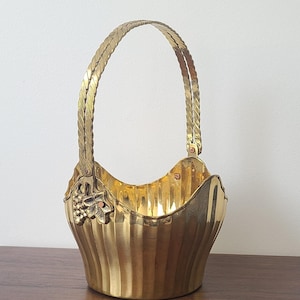 May include: A polished brass basket featuring a vertical fluted design and a single braided handle. The basket is adorned with a decorative metal grape cluster and leaf motif on each side where the handle attaches to the scalloped rim.