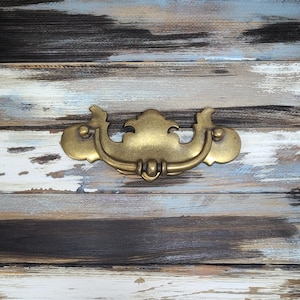 Peut inclure: Une poignée de tiroir dorée avec un design décoratif. La poignée est montée sur une surface en bois vieilli avec une finition de peinture bleue et brune.