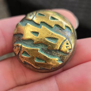 May include: A round metal button featuring a raised relief design of three swimming fish. The surface has a polished gold-tone finish on the fish figures, contrasted by a recessed, oxidised teal-green patina in the background areas of the button.