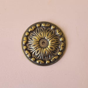 May include: A gold-toned metal decorative rosette with a floral design. The rosette has a small hole in the centre.