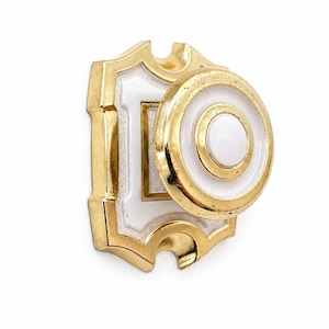 May include: A decorative door knob with a white center and gold trim. The knob has a circular design with concentric rings and is mounted on a white and gold escutcheon plate. The hardware is likely made of metal.