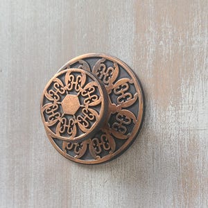 May include: A round cabinet knob featuring a copper-colored metal face with a raised, intricate floral-like pattern and a central hexagon. The design is set against a dark, recessed background, creating a layered, circular appearance on a surface.
