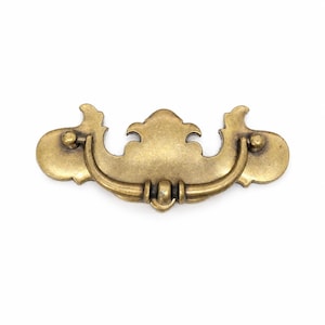 May include: A brass-toned drawer pull with an ornate design. The pull features a curved handle and decorative backplates with a symmetrical, vintage aesthetic. The hardware is likely for cabinets or furniture, adding a classic touch.