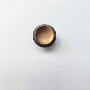 May include: A circular knob featuring a matte black outer rim and a recessed, metallic gold-toned centre. The interior of the gold-coloured basin displays a subtle, textured finish with a small, faint marking visible near the upper edge.
