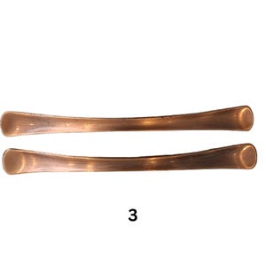 May include: Two curved, metallic copper-toned cabinet pulls with a smooth, elongated design and rounded ends. Each handle features a polished finish with subtle variations in colour. The number 3 is printed in black below the two handles.