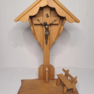 Peut inclure: Une petite église miniature en bois avec un toit, une croix avec une figurine de Jésus et un petit banc. L'église est en bois brun clair et a un aspect rustique.